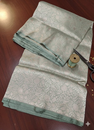 Mint green Banarasi saree fabric with intricate zari floral weaving, neatly folded with a soft sheen, placed alongside a thread spool and scissors on a wooden surface.